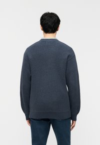 Άνδρας που φοράει σκούρο μπλε πλεκτή μπλούζα και τζιν, εμφανιζόμενος από πίσω με φόντο ένα απλό λευκό φόντο.