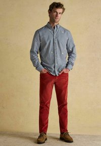 Modrá a biela pruhovaná košeľa s gombíkmi a farebnou výšivkou, v kombinácii s červenými kordovými nohavicami a hnedými topánkami, stojí pred neutrálnym pozadím.