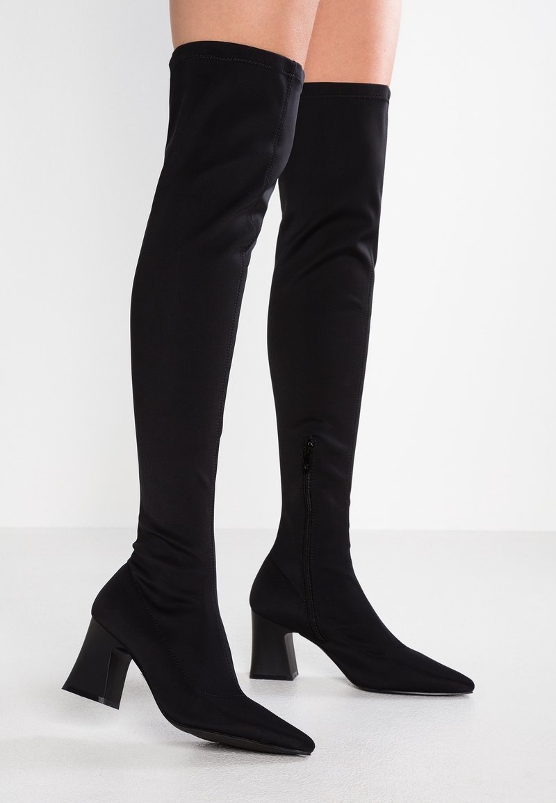 Bottes noires mi-cuisses en tissu extensible avec un bout pointu. Elles présentent une surface texturée et un talon carré épais pour plus de stabilité.