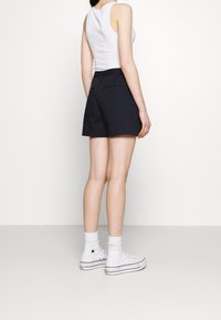 Pantalones cortos negros de talle alto con bolsillos traseros, combinados con una camiseta sin mangas blanca y zapatillas blancas de caña alta. Tejido suave, diseño casual.