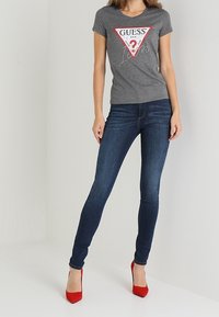 Grå bomullströja med "GUESS"-logotyp i en röd triangel, ihop med mörkblå skinny jeans och röda högklackade skor.