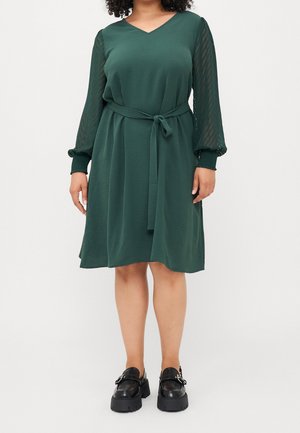 Frau trägt ein dunkelgrünes knielanges Kleid mit durchsichtigen, strukturierten langen Ärmeln und einem Stoffgürtel, kombiniert mit schwarzen klobigen Plateau-Schuhen.