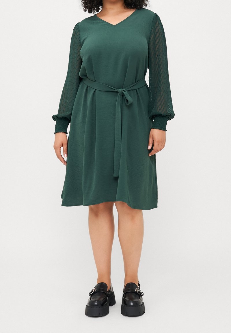 Femme portant une robe vert foncé mi-longue avec des manches longues transparentes texturées et une ceinture en tissu, associée à des chaussures plateformes noires épaisses.