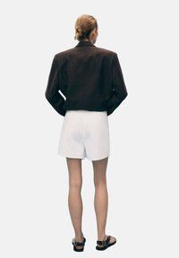 Veste courte marron, shorts blancs et sandales noires. Textures lisses avec une coupe slim. Manches courtes et silhouette épurée.