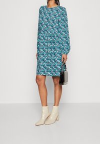 Blommönstrad klänning i teal och blått, långärmad, rund hals, avslappnad passform, kombinerad med glänsande beige ankelboots och en liten svart handväska.