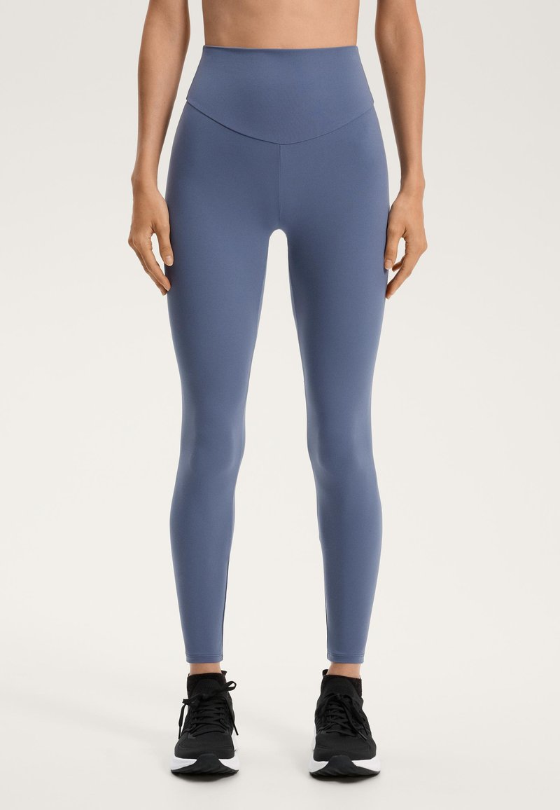 High-waisted blue leggings made from smooth, stretchy fabric. Features a seamless design without noticeable patterns. Black athletic shoes.