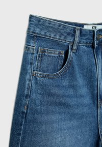 Close-up van een blauwe denim jeans taille met voor- en muntzakken, riemlussen en een metalen knoopsluiting.