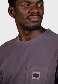 Fialové bavlnené tričko s vreckom a čiernym logom slona. Tričko má okrúhly výstrih a viditeľné švy.