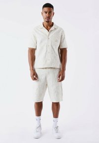 Camisa de botones de manga corta y pantalones cortos a juego en tela beige clara con patrón texturizado. Zapatillas blancas y calcetines tipo crew completan el conjunto.