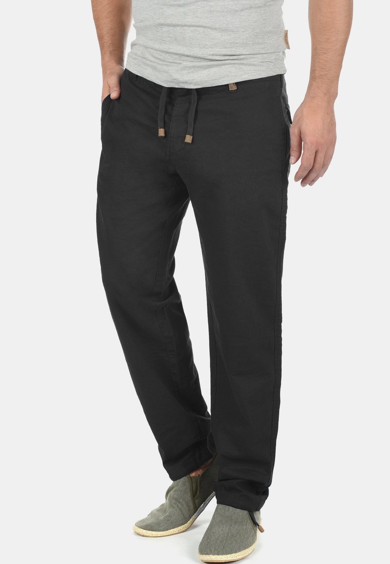 Pantalones de algodón negros con un corte holgado, que cuentan con una cintura ajustable con cordón y bolsillos laterales. Combinados con zapatos casuales verdes sin cordones.