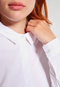 Vit skjorta med klassisk krage, glänsande knappar och mjuk tygtextur, som visar manschettdetaljer och en ren, skräddarsydd look.