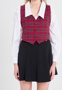 Czerwona i zielona kamizelka w kratę z ostrym białym kołnierzykiem, z zapięciem na guziki i lekko skośnym dołem, zestawiona z prostą czarną spódnicą.