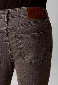 Κοντινό πλάνο ατόμου που φοράει σκούρα γκρι τζιν denim με πίσω τσέπη και καφέ δερμάτινο λογότυπο Calvin Klein στη μέση.