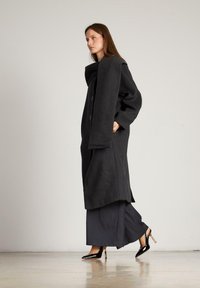 Largo abrigo negro de lana con un collar oversize, botones en el frente y bolsillos laterales, combinado con pantalones oscuros de pierna ancha y tacones de charol puntiagudos.