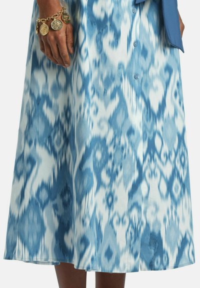 Ruka s náramkom s príveskom zlatých mincí spočíva vedľa sukne strednej dĺžky s modro-bielym abstraktným kvetinovým vzorom a gombíkovými detailmi.