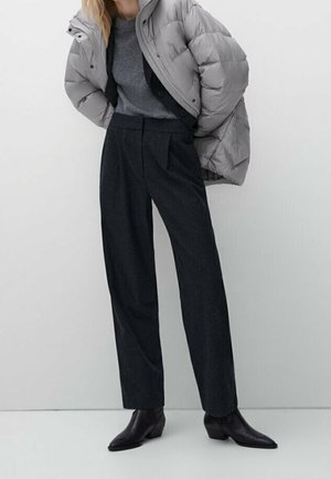 Femme portant une doudoune grise par-dessus un pull gris, un pantalon large taille haute noir et des bottines noires, debout devant un fond uni.