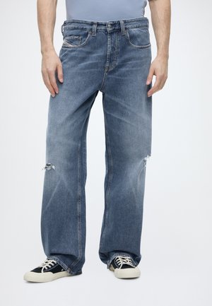 Homme portant un jean bleu ample avec de petites déchirures aux deux genoux et des baskets noires et blanches sur un fond uni.