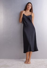 Vestido de satén negro, tipo slip, con tirantes finos, dobladillo a la altura de la rodilla, textura suave y un corte relajado, lucido por una modelo que está de pie sobre una superficie suave.