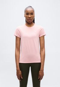 Γυναίκα που στέκεται όρθια, κοιτώντας μπροστά, φορώντας ένα ανοιχτό ροζ μπλουζάκι Puma και σκούρο ελιά παντελόνι, με φόντο καθαρό λευκό.