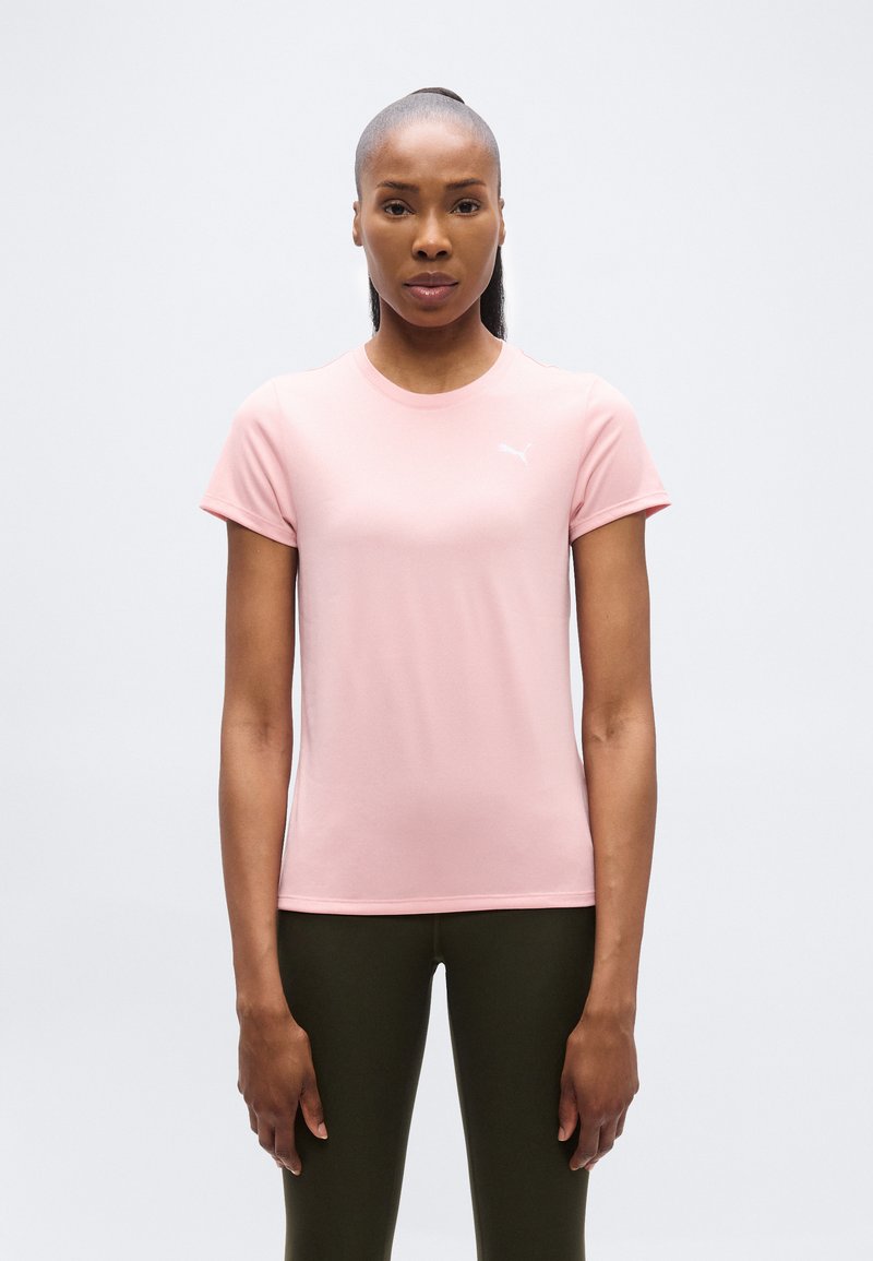 Γυναίκα που στέκεται όρθια, κοιτώντας μπροστά, φορώντας ένα ανοιχτό ροζ μπλουζάκι Puma και σκούρο ελιά παντελόνι, με φόντο καθαρό λευκό.