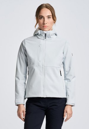 Una mujer luce una chaqueta gris claro con cremallera, cuello alto y capucha, combinada con unos pantalones oscuros sobre un fondo liso.