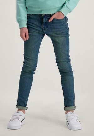 Strakke blauwe jeans met een medium wassing en een taille die net onder de navel valt. Draagt witte sneakers en heeft een opgerolde zoom bij de enkels.
