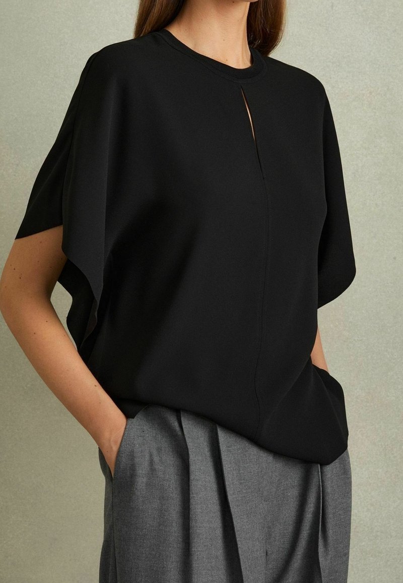 Femme portant un haut noir ample à manches courtes avec une petite fente à l'avant, associé à un pantalon plissé gris taille haute, mains dans les poches.