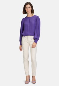 Lilla bluse med lange ærmer og samlet halsudskæring, parret med tætsiddende cremefarvede bukser. Modellen bærer lavendelfarvede højhælede sandaler.