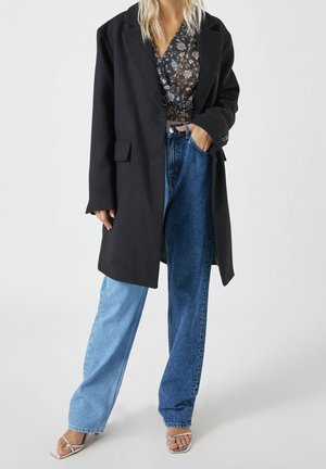 Schwarzer oversized Mantel mit umgeschlagenem Revers, getragen über einem floral gemusterten Wickeloberteil, kombiniert mit hellblauen und dunkelblauen weiten Jeans und silbernen High Heels.