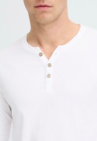 Bijela dugorukava henley majica s okruglim izrezom, plaketom od tri drvene gumba i mekanim, teksturiranim materijalom.