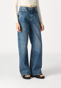 Mėlyno džinsinio audinio plačių kojų kelnės su aukšta talija, penkių kišenių dizainu ir šiek tiek pažeidimų klubų srityje. Suderintos su juodais platforminiais sandalais.