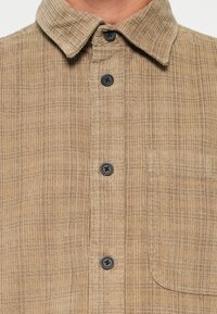Bruine corduroy overhemd met een button-down kraag, zwarte knopen en een borstzak met een gestructureerd, gestreept patroon.