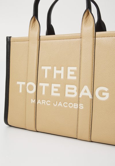 Beige leather tote bag with black accents, featuring prominent white text "THE TOTE BAG" and "MARC JACOBS" on the front. Smooth texture.