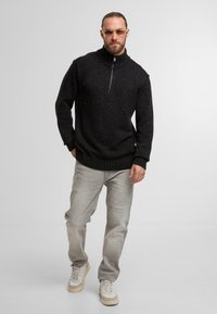 Schwarzer Pullover mit hohem Kragen und Reißverschluss, ausgestattet mit strukturiertem Strick und gesprenkeltem Muster, kombiniert mit hellgrauen Jeans und Sneakers.