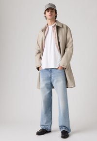 Νεαρός άνδρας με μπεζ καπαρντίνα, λευκό μπλουζάκι, ανοιχτό μπλε τζιν, μαύρα παπούτσια και γκρι καπέλο, στέκεται με τα χέρια στις τσέπες.