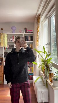 Černá fleecová zipová mikina s vysokým límcem, doplněná červenými kostkovanými kalhotami. Stojící před policí s knihami a pokojovými rostlinami.
