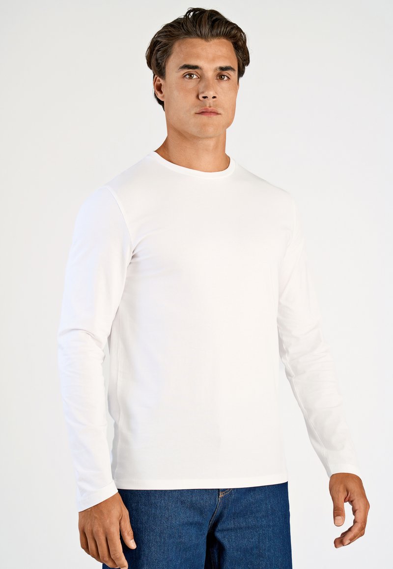 Valkoinen pitkähihaisen t-paita, joka on tehty pehmeästä puuvillasta. Paidassa on pyöreä kaula-aukko ja se on vartalonmyötäinen, käytetty sinisten farkkujen kanssa.