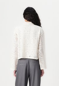 Biała szydełkowana krótka kardiganka z kwiatowymi wzorami w wycięciach i długimi rękawami, w połączeniu z szarymi spodniami z wysokim stanem.