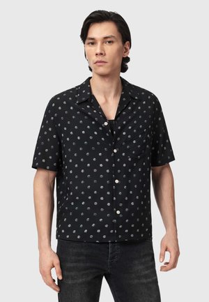 Młody mężczyzna w czarnej koszuli z krótkim rękawem w białe grochy oraz ciemnych dżinsach, stojący na jednolitym tle.