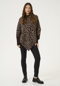 Ruskea ja beige leopardikuosisweater, jossa on timanttimallinen helma, tekstuuri neulottu ja roikkuneet olkapäät; yhdistetty mustiin slim-fit-housuihin ja mustiin kenkiin.