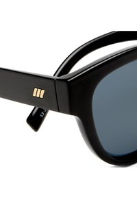 Κοντινό πλάνο μαύρων γυαλιών ηλίου με παχιά σκελετό και σκοτεινούς φακούς, με μια μικρή χρυσή λεπτομέρεια τριπλής μπάρας στην πλαϊνή πλευρά.