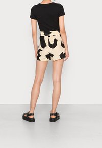 Black short-sleeve top with scalloped edges, paired with cream shorts featuring large black abstract patterns. Model wearing black sandals.