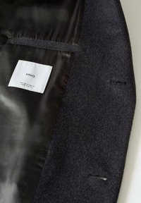 Manteau en laine noir avec doublure satinée noire, présentant une étiquette blanche indiquant "MNG" et les détails de la taille à l'intérieur du manteau.