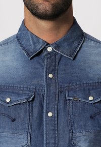 Denimskjorta i ljusblå nyans, med spetsig krage, knappstängning framtill, två bröstfickor och synliga sömnadsdetaljer.