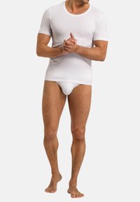 Mann in einem engen weißen kurzärmeligen Hemd und passenden weißen Unterhosen, barfuß stehend mit gefalteten Händen vor sich.