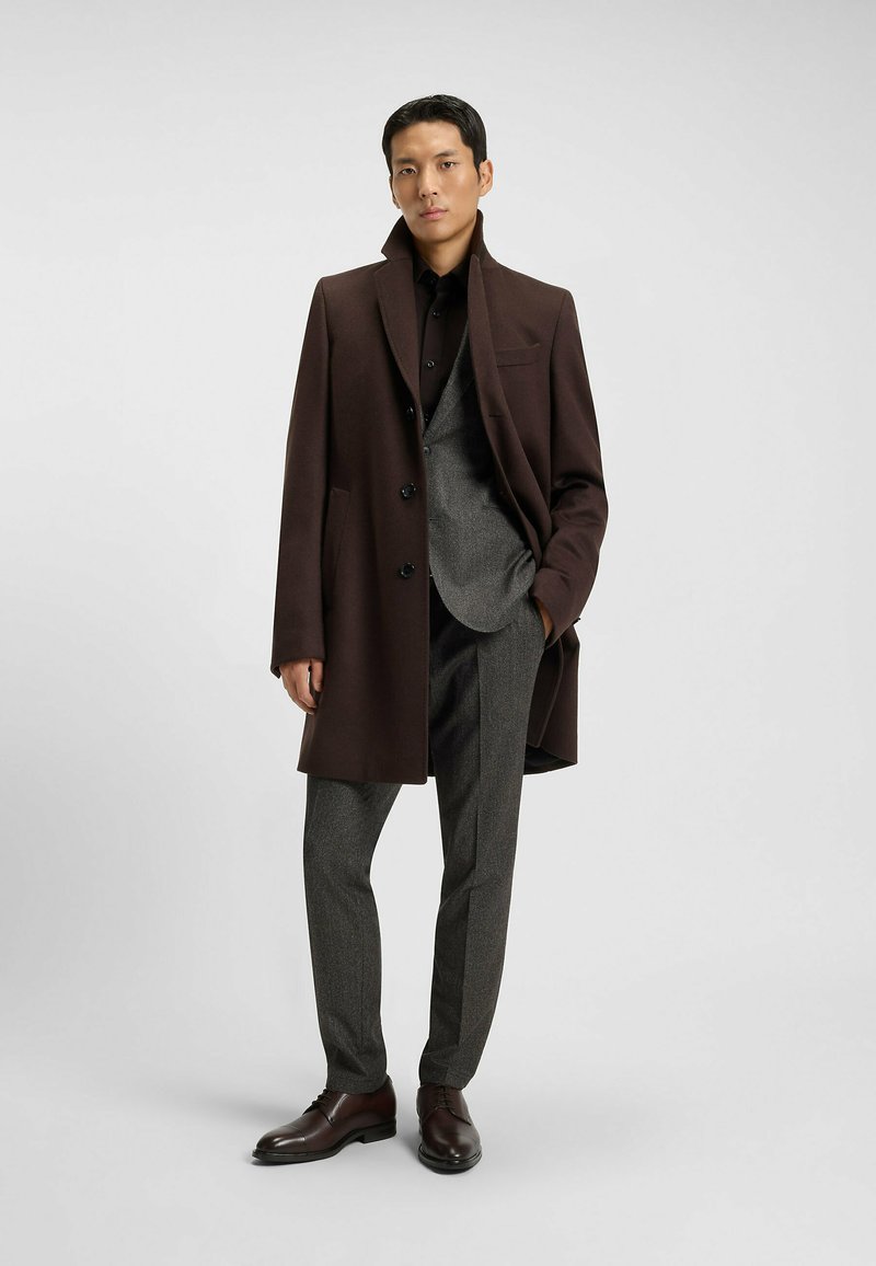 Cappotto di lana marrone con un colletto strutturato, abbinato a un gilet grigio antracite, pantaloni slim e scarpe in pelle scura. Texture liscia e vestibilità su misura.