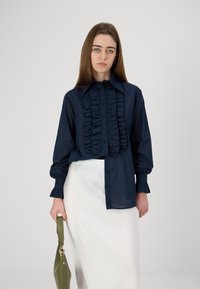 Cămășă bleumarin cu față volan, cu închidere prin buton, având mâneci lungi cu manșete volan, combinată cu o fustă albă drapată și o geantă verde.