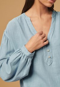 Lichtblauwe linnen blouse met een V-hals, drie knopen bij de halslijn en aangeknoopte mouwen. Gladde textuur met een relaxte pasvorm.