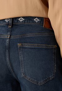 Mørkeblå denimjeans har en baklomme med synlige sømmer og en tan lærlapp. En beigefarget stoffoverdel dekker delvis linningen.