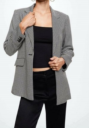 Kvinde iført en sort tube top, sorte bukser og en sort-hvid hundetandsmønstret blazer, justerer blazeren revers med den ene hånd.